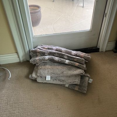 Sale Photo Thumbnail #141: Stack of plush, grey tufted bath mats and accent rugs folded on the carpet inside a sliding glass door.