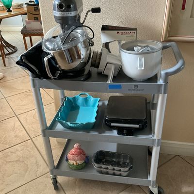 Sale Photo Thumbnail #96: A gray three-tier utility rolling cart filled with various kitchen small appliances and bakeware, including a silver KitchenAid bowl-lift stand mixer with attachments, a blue baking dish, and a waffle maker.