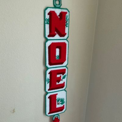 Sale Photo Thumbnail #131: Handmade plastic canvas "NOEL" wall hanging with red stitched lettering, holly accents, and a yarn tassel bottom.