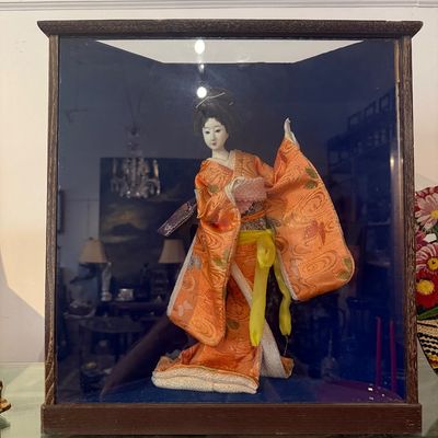Vintage Japanese geisha doll wearing an ornate orange silk kimono, housed in a wooden glass display case.