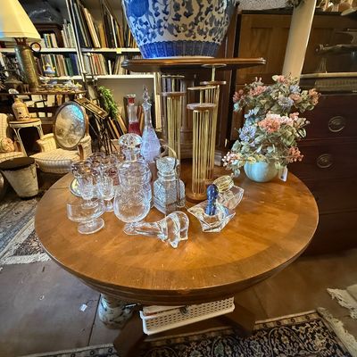 Collection of vintage cut glass decanters, glassware, brass candlestick holders, and home decor arranged on a round wooden table.