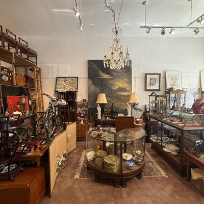 Antique and vintage estate sale showroom featuring a crystal chandelier, curved glass display cases filled with collectibles, framed...