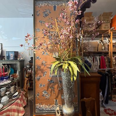 Tall decorative silver-tone embossed metal vase featuring a large artificial pink orchid arrangement, displayed alongside a framed...