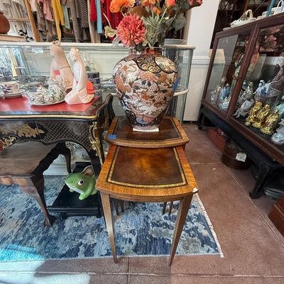 Pair of vintage nesting tables with leather-insert tops, displayed alongside a large ornate Asian porcelain vase holding faux floral...