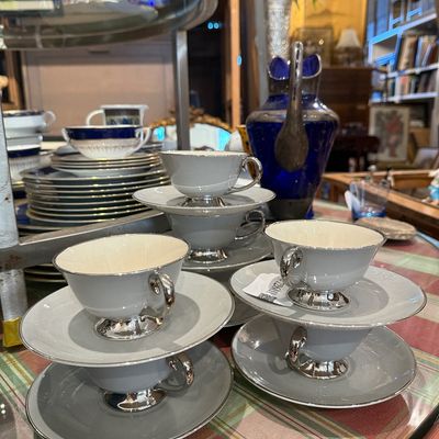 Set of grey porcelain teacups and saucers with silver-plated trim and handles, displayed alongside a cobalt blue glass pitcher.