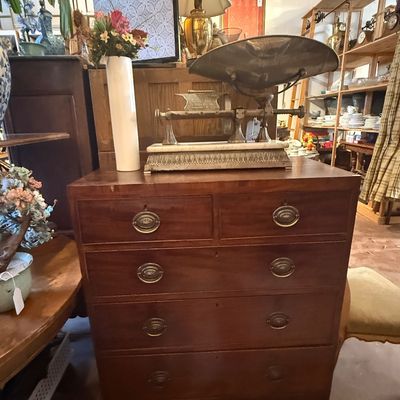 Antique mahogany chest of drawers with brass oval pull handles, topped with a vintage mechanical scale and a decorative floral vase.