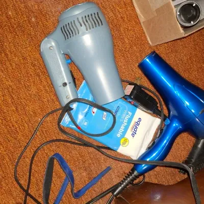 Sale Photo Thumbnail #143: Collection of personal care items including a metallic blue hair dryer, a grey compact hair dryer, and a pack of Equate flushable wipes resting on carpeting.