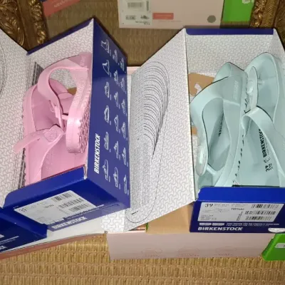 Selection of boxed EVA sandals including pink and light blue Birkenstock EVA thongs alongside a pair of tie-dye Crocs slide sandals, all...