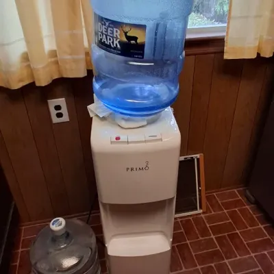 White Primo top-loading water dispenser with a 5-gallon Deer Park water bottle attached, accompanied by an extra water jug.