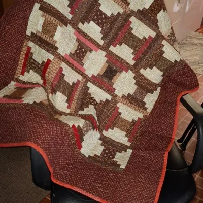 Handmade patchwork log cabin pattern quilt draped over an office chair, featuring earth tones of brown, cream, and red accents.
