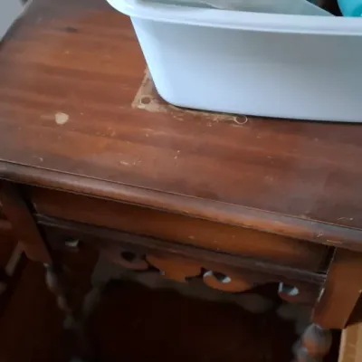 Vintage dark stain wooden side table with carved apron and turned legs, featuring surface wear and holding a white plastic tub of novelty...