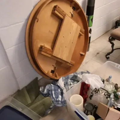 Sale Photo Thumbnail #195: Round wooden table top leaning against a white block wall, surrounded by plastic storage totes filled with household items.