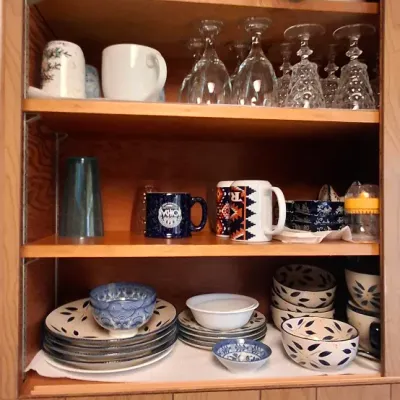 Open wooden kitchen cabinet filled with assorted glassware, coffee mugs, Pyrex casserole dishes, and patterned ceramic bowls.