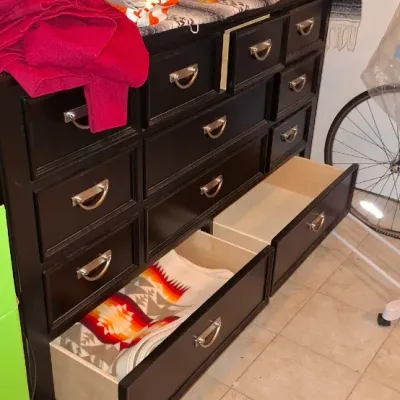 Sale Photo Thumbnail #222: Large black multi-drawer dresser featuring silver bail handles, partially filled with linens and displaying decorative figurines and a mirror on top.