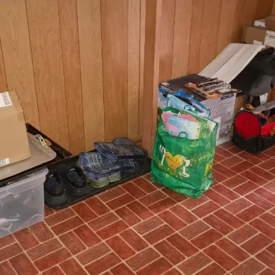 Assorted household items lined up against a wood-paneled wall, including a clear storage tub, silver trays, cardboard boxes, shoes,...