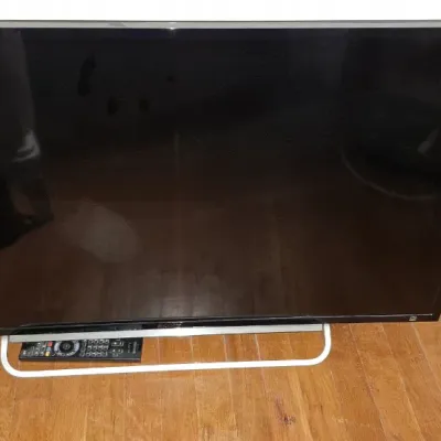 Sale Photo Thumbnail #267: Black flat-screen television with a silver metallic loop stand, resting on a hardwood floor alongside a remote control.