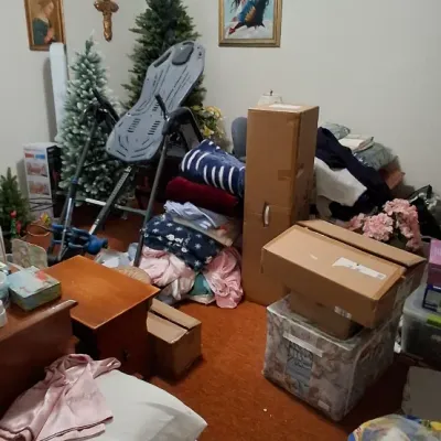 Room filled with assorted household items, including Teeter inversion table, artificial Christmas trees, framed wall art, bedding stacks,...