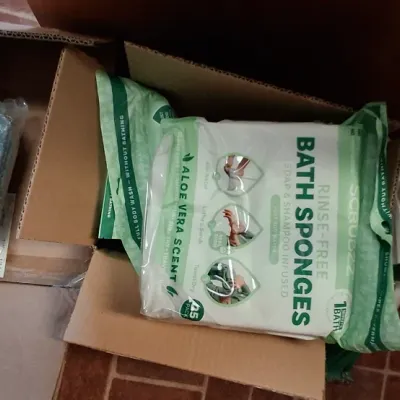 Cardboard boxes filled with personal care and household items, including a package of Scrubzz rinse-free bath sponges and a rolled light...