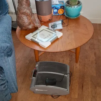 Sale Photo Thumbnail #254: Dr. Ho's Motionciser foot massager on the floor next to a round mid-century style wooden side table holding a carved bear statue, humidifier, potted cactus, and decor.