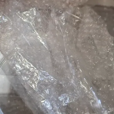 Clear glass or acrylic lid partially wrapped in protective bubble wrap inside a cardboard storage box.