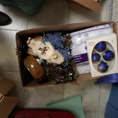Sale Photo Thumbnail #171: Cardboard box filled with mixed Christmas decorations, including blue glass baubles, metallic tinsel garland, a fur-trimmed figurine, and glittery snowflake ornaments.