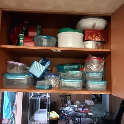 Wooden upper kitchen cabinet packed with assortment of plastic food storage containers and decorative tins, over a counter displaying...