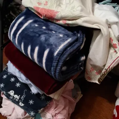 Sale Photo Thumbnail #136: Stack of assorted household linens and blankets, including a blue plush throw, maroon blanket, snowflake-patterned fleece, and embroidered textiles.