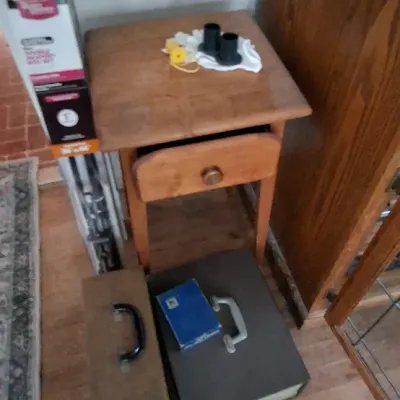 Sale Photo Thumbnail #246: Small wooden single-drawer nightstand or side table, shown with vintage carrying cases and household hardware on the floor.
