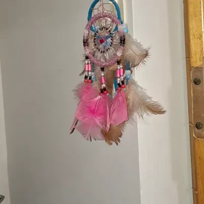 Sale Photo Thumbnail #116: Handcrafted dreamcatcher featuring pink and blue beaded web designs with dangling pink and light brown feathers, hanging from a plastic wall hook.