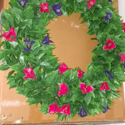 Artificial green foliage wreath decorated with bright pink and purple flowers, displayed atop a cardboard shipping box.