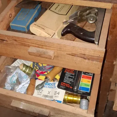 Sale Photo Thumbnail #228: Open wooden workshop drawers containing various vintage tools and hardware items, including hand planes, saw filing guides, and packaged supplies.