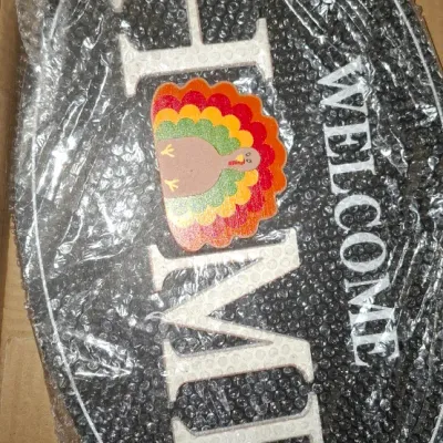 Oval-shaped "Welcome Home" decorative plaque wrapped in bubble wrap, featuring white lettering and a colorful Thanksgiving...