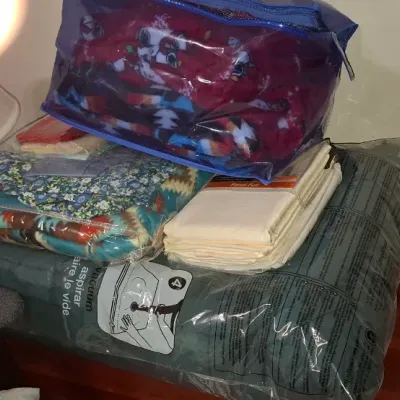 Assortment of packaged linens and blankets, including a vacuum-sealed storage bag of bedding, folded cream sheets, patterned fleece...