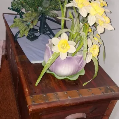 Sale Photo Thumbnail #132: Rustic wood trunk with metal stud trim and side handles, topped with artificial daffodils in a purple ceramic pot next to a pre-lit artificial Christmas tree base.