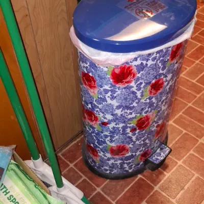 The Pioneer Woman floral-printed stainless steel step trash can, featuring a blue lid, shown alongside a green broom and household...