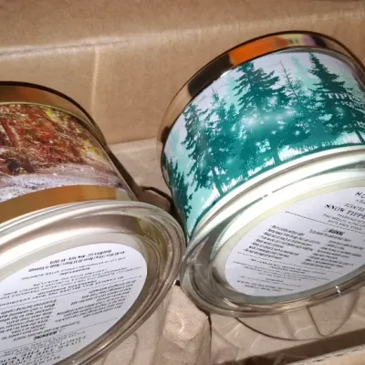 Two Homeworx by Slatkin & Co. scented 3-wick candles, including "Snow Tipped Pine", sitting inside cardboard shipping...