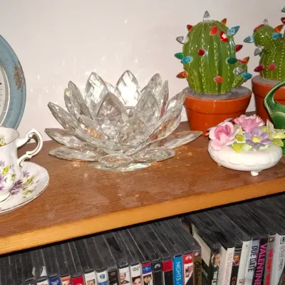 Sale Photo Thumbnail #272: Collection of decorative home accents, including a rustic light blue wall clock, a glass lotus candle holder, lighted ceramic cactus figurines, and porcelain floral knick-knacks arranged on a wooden shelf above DVDs.
