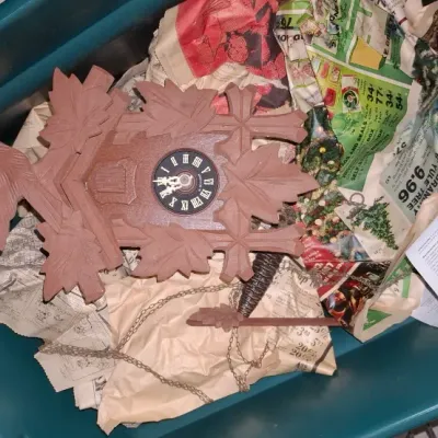 Sale Photo Thumbnail #173: Vintage carved wooden cuckoo clock with bird and leaf motifs, packed inside a plastic storage tote with newspaper.