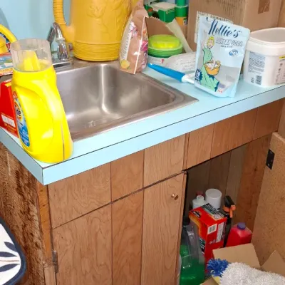 Vintage utility sink cabinet and laundry area filled with assorted cleaning supplies, a yellow watering can, and household items.