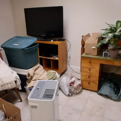 Sale Photo Thumbnail #215: Collection of household items including a wooden desk, flat-screen TV on a media console, white portable dehumidifier, and a wooden armchair with throw blankets.