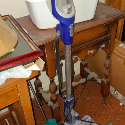 Shark modern blue corded stick vacuum cleaner with attachments, shown standing next to wooden furniture.