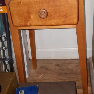 Sale Photo Thumbnail #248: Vintage light wooden nightstand with a single drawer and tapered legs, shown with a vintage Argus film strip adapter in its original box sitting on top of a carrying case in the foreground.