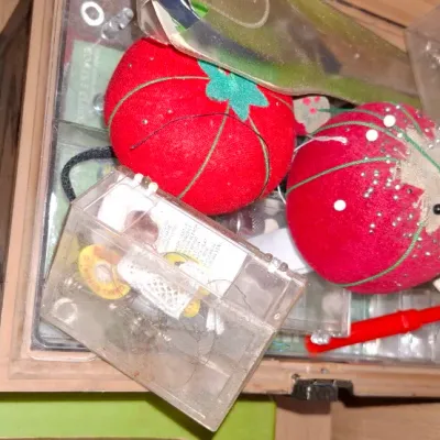 Collection of sewing supplies inside a drawer, including red classic tomato pincushions, plastic accessory cases, bobbins, and a seam...