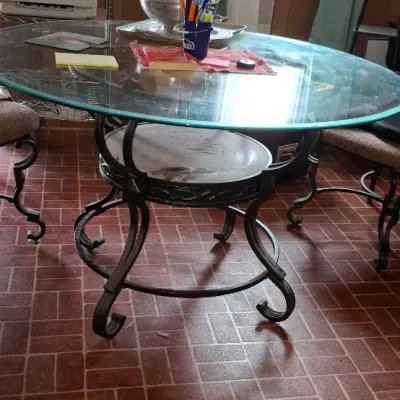 Round glass-top dining table featuring a wrought iron base with scroll legs, accompanied by matching upholstered chairs.
