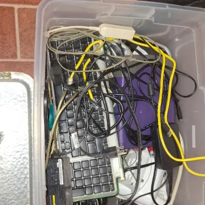 Clear plastic storage tote filled with assorted electronics, including computer keyboards, power bricks, ethernet cables, and small...
