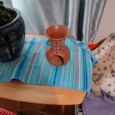Sale Photo Thumbnail #126: Terracotta sun face essential oil burner resting on a blue striped table runner alongside a glazed ceramic potted plant.