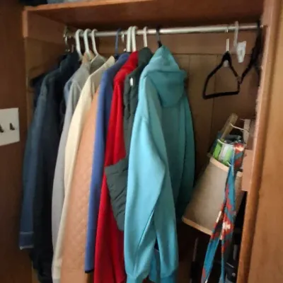 Wooden storage closet filled with hanging outerwear, plastic storage bins, woven baskets, and miscellaneous household items.