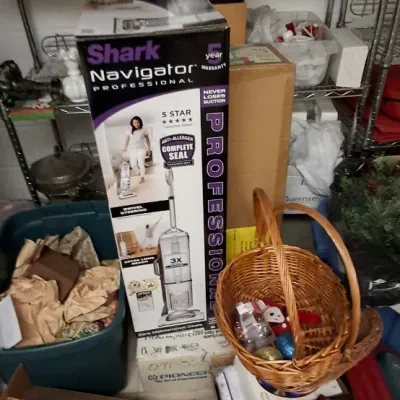 Sale Photo Thumbnail #168: Shark Navigator Professional upright vacuum boxed alongside wicker baskets, plastic storage totes, and miscellaneous holiday decor items on wire shelving.