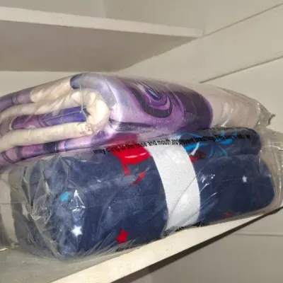 Two soft plush fleece throw blankets sealed in original plastic packaging, stacked on a closet shelf.