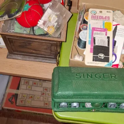 Collection of vintage sewing supplies, including a green Singer attachment case, tomato pincushions, thread spools, and packaged hand...